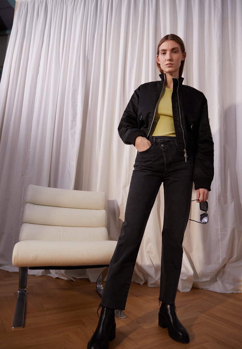 Black bomber jacket over a lime green ribbed top, paired with dark straight-leg jeans and black ankle boots; stands beside a white chair.