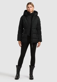 Kvinde iført en sort polstret hættetrøje, sorte leggings og sorte støvler til midten af læggen, der står foran en ensfarvet baggrund.