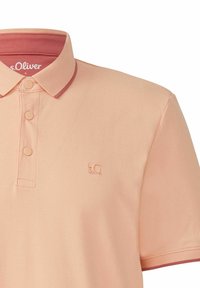 Helles Pfirsich-Polohemd mit einem kontrastierenden burgunderfarbenen Kragen. Besitzt drei Knöpfe und ein kleines, geprägtes Logo auf der Brust. Hergestellt aus strukturiertem Stoff.
