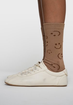 Weiße, strukturierte Leder-Sneaker mit niedrigem Profil, kombiniert mit braunen, gerippten Socken, die ein sich wiederholendes Muster von lächelnden Emojis zeigen.