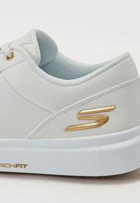 White leather sneaker with a gold metallic logo, padded collar, and textured tongue. Features a light blue interior and cushioned sole.