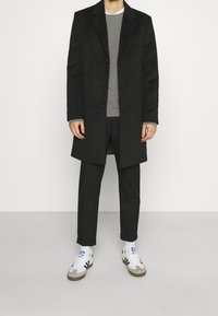 Schwarzer Wollmantel mit drei Knöpfen, grauer Pullover darunter, schwarze Hose und weiße Sneakers mit schwarzen und braunen Akzenten.