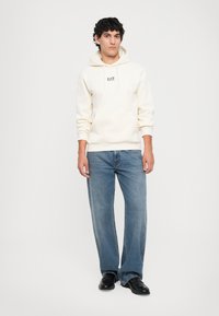 Ung mann iført en kremfarget EA7-hettegenser, blå jeans og svarte sko stående mot en enkel hvit bakgrunn.