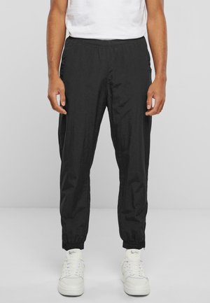 Person trägt schwarze elastische Bundhose und weiße Sneakers, steht vor einem einfarbigen hellgrauen Hintergrund.