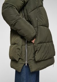 Chaqueta acolchada en verde olivo oscuro con un diseño acolchado, bolsillos laterales con cremallera y puños de canalé. El material parece suave y aislante.