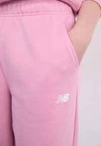 Pantalon de survêtement rose en tissu doux avec une taille élastique et des poches latérales. Présente un logo New Balance brodé en blanc sur la cuisse.