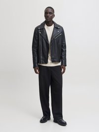 Mann som står iført svart skinnjakke over en beige genser, svarte vide bukser og svarte dressko mot en enkel bakgrunn.