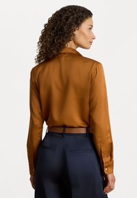 Καφέ σατινένιο πουκάμισο με μακριά μανίκια και γιακά, σχεδιασμένο με απαλή υφή. Συνδυασμένο με ναυτικά παντελόνια που έχουν δερμάτινη ζώνη.