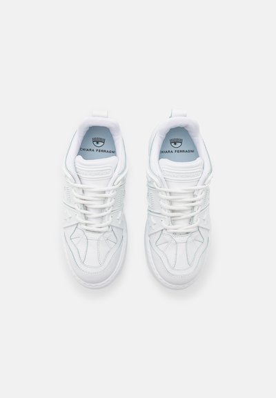 Baltās sporta kedas ar gludu ādas virsu, kontrastējošu diegu šuvēm, zvaigžņu formas akcentiem un plakanu zoli ar auklēm.