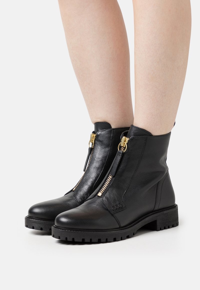Bottines en cuir noir avec semelles épaisses et fermetures éclair dorées à l'avant, portées sur des jambes nues sur un fond clair uni.