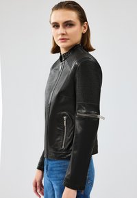 Chaqueta de cuero negro con acentos acanalados en los hombros, bolsillos con cremallera y un cuello alto. Presenta un diseño ajustado y una textura suave.