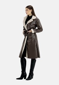 Brauner Kunstlederlanger Mantel mit cremefarbenem Kunstlammfellkragen, tailliert gebunden, Seitentaschen und glatter Textur. Kombiniert mit schwarzen knielangen Stiefeln.