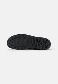 Semelle de botte en caoutchouc noir avec des crampons profonds comprenant des chevrons, des barres et des formes de losange, présentée sur un fond blanc uni.