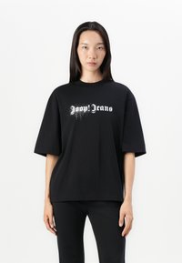 T-shirt noir surdimensionné en coton, avec un design graphique de texte blanc "Jeans" avec un effet de éclaboussure sur le devant, manches courtes.