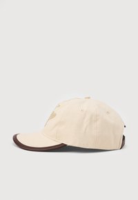Beige cotton baseball cap with brown leather-trimmed brim and adjustable strap, shown in side profile on a plain background.