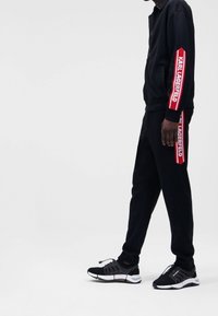 Chándal negro con rayas rojas y el texto "KARL LAGERFELD" en las mangas y los laterales. Combinado con zapatillas deportivas en blanco y negro.