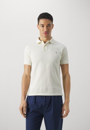 Light cream short-sleeve polo shirt with collar, featuring a small embroidered logo. Worn with navy blue trousers, displaying a casual look.