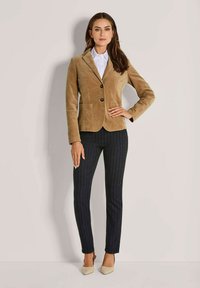 Frau steht mit der Hand auf der Hüfte, trägt einen hellbraunen Cordblazer, ein weißes Hemd, eine dunkel gestreifte Hose und beigefarbene spitze Pumps.