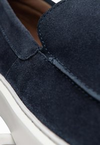 Gros plan d'une chaussure en daim bleu marine à enfiler avec des coutures détaillées et une semelle en caoutchouc blanche.