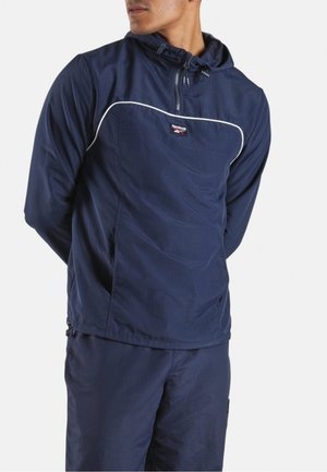 Man in een marineblauw Reebok trainingspak met ritskap en witte biezen, staand met de handen achter de rug, licht naar rechts gericht.
