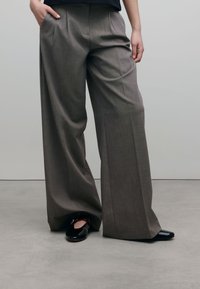 Grijze wijde broek met een gestructureerde weefsel en voorplooien, gecombineerd met zwarte glanzende schoenen. De stof lijkt gestructureerd en op maat gemaakt.