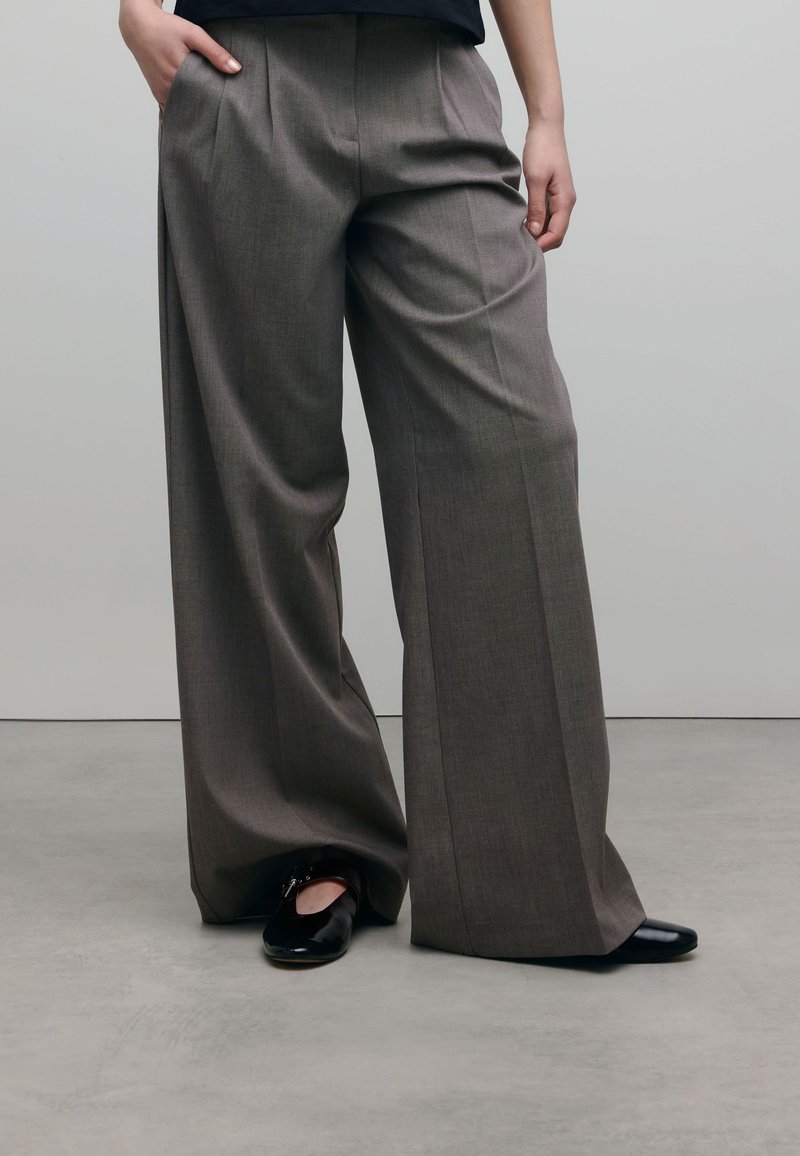 Grijze wijde broek met een gestructureerde weefsel en voorplooien, gecombineerd met zwarte glanzende schoenen. De stof lijkt gestructureerd en op maat gemaakt.