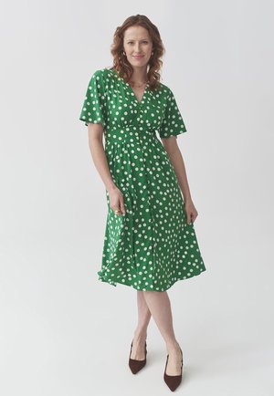 Robe verte à pois blancs, manches courtes, devant à boutons et jupe évasée. Portée avec des escarpins marron à bout pointu.