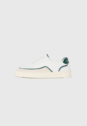 Zapatilla baja blanca con detalles en verde, gamuza beige y suela de color crema con un fondo estriado.