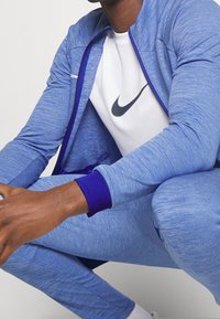 Blå atletisk jakke med lilla detaljer og lynlåslukning, kombineret med en hvid Nike t-shirt. Stoffet har et meleret udseende.
