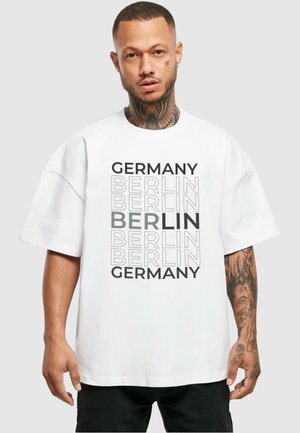 Hvit oversized t-skjorte med svart tekst som leser "Tyskland" og "Berlin" i varierte størrelser og tykkelser, rund hals, korte ermer.
