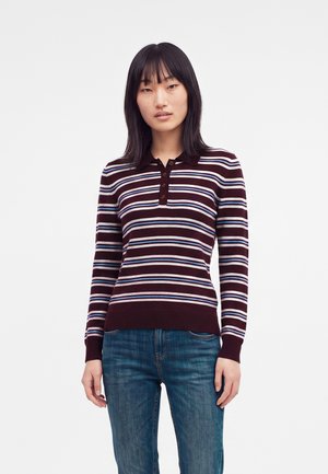 Jeune femme aux cheveux noirs et raides portant un polo à manches longues rayé marron, blanc et bleu et un jean bleu sur un fond uni.