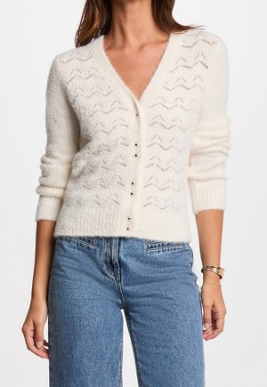 Femme portant un cardigan en maille crème boutonné avec des motifs en zigzag et des manches retroussées, associé à un jean bleu taille haute.
