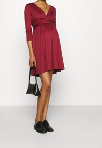 Vinröd knälång klänning med djup V-ringning, vridningsdetalj vid midjan, trekvartsärmar, tillsammans med en liten svart handväska och skor.