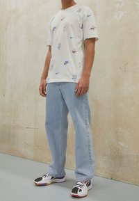 Camiseta blanca con logos de Nike repetidos en azul, rosa y púrpura. Llevada con jeans de mezclilla azul claro y zapatillas multicolores.