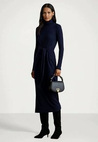 Femeie îmbrăcată cu o rochie midi cu mâneci lungi și guler înalt de culoare bleumarin, cu cizme negre cu toc și ținând în mână o geantă mică neagră cu un închizător auriu.