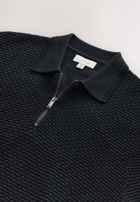 Črn pulover z zadrgo teksturirane tkanine, ovratnik z ovratnikom in rebrastimi detajli, z srebrno zadrgo in značko z napisom "NEXT SIGNATURE".