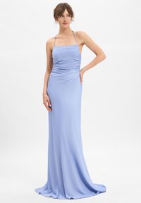 Femme portant une robe satinée longue et légère de couleur bleu clair, avec des bretelles fines, un corsage froncé et une légère traîne, se tenant contre un fond blanc.
