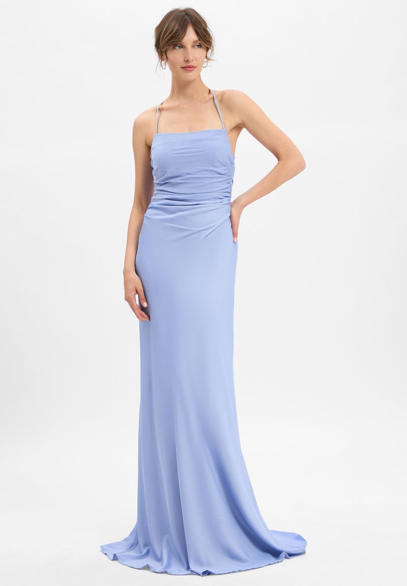 Femme portant une robe satinée longue et légère de couleur bleu clair, avec des bretelles fines, un corsage froncé et une légère traîne, se tenant contre un fond blanc.