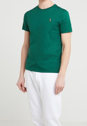 Grünes Baumwoll-T-Shirt mit Rundhalsausschnitt, kurzen Ärmeln und einem kleinen gestickten Logo auf der Brust, kombiniert mit weißen Hosen.