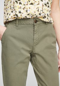 Persona che indossa pantaloni verde oliva con tasche e una camicetta floreale infilata dentro, con motivi gialli, marroni e viola.