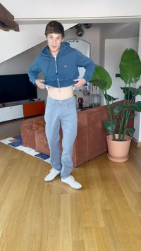 Hanorac albastru scurt, pantaloni albastru deschis și adidași albi. Stând pe podea de lemn, lângă o canapea maro și o plantă mare verde.