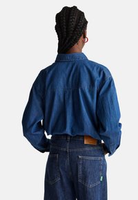 Camicia di jeans blu con vestibilità rilassata, colletto button-down e maniche lunghe. Presenta un dettaglio sulla spalla e cuciture a vista, abbinata a jeans scuri.