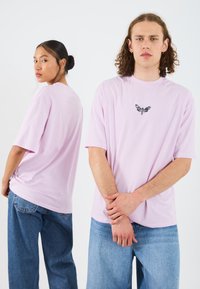 Light pink short-sleeve T-shirt featuring a black butterfly graphic on the chest, paired with light blue jeans. Smooth fabric, relaxed fit.