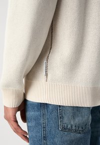 Jersey de punto crema con ribetes en el bajo y los puños, que presenta un zipper lateral decorativo y una etiqueta de marca cerca de la costura. Lleva puesto sobre denim.