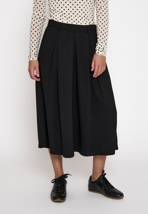 Kvinde iført en hvid langærmet bluse med polkaprikker, sort plisseret midi-nederdel og sorte snørebåndssneakers, der står mod en hvid baggrund.
