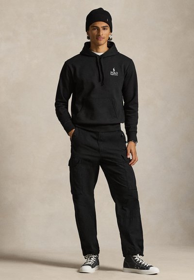 Fiatal férfi fekete sapkában, fekete Polo Ralph Lauren kapucnis pulóverben, fekete katonai nadrágban, és fekete magas szárú tornacipőben fehér fűzővel és talppal.