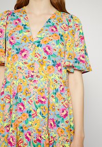 Vestido floral com decote em V, apresentando um fundo azul e flores vibrantes em rosa, laranja e amarelo. Mangas curtas com babados e tecido texturizado.