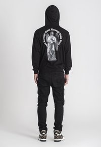 Sudadera negra con un gran gráfico de un ángel en la parte posterior, acompañado de texto alrededor. Combinada con pantalones cargo negros y zapatillas con un diseño estampado.