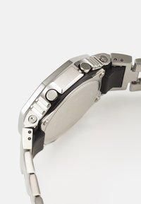 Montre en acier inoxydable argenté avec un bracelet en caoutchouc noir. Présente un cadran plat et poli ainsi que plusieurs boutons sur le côté pour des fonctionnalités.