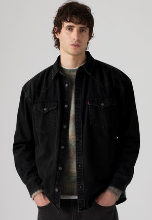 Jeune homme aux cheveux bouclés foncés portant une veste en jean noire boutonnée sur un pull multicolore, les mains dans les poches, fond neutre.